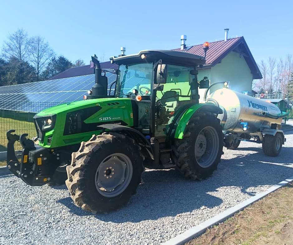 Zielony traktor marki Deutz-Fahr model 5105 Keyline stoi na żwirowej drodze, ciągnąc za sobą metalową beczkowóz marki Meprozet. W tle widoczny jest budynek z ciemnoczerwonym dachem oraz duża instalacja paneli fotowoltaicznych. Pogoda jest słoneczna, a cienie maszyn są dobrze widoczne na podłożu.