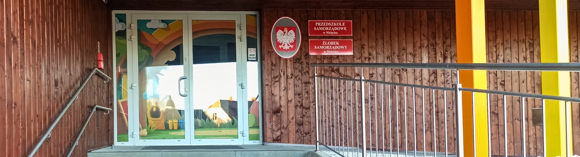 Wejście do budynku przedszkola i żłobka samorządowego w Niebylcu. Po lewej stronie znajdują się białe drzwi z kolorową, dziecięcą grafiką przedstawiającą krajobraz z tęczą, domkiem i drzewami. Obok drzwi umieszczone są dwie czerwone tablice z nazwami placówek oraz godło Polski. Budynek wykończony jest drewnianą elewacją, a po prawej stronie widoczne są kolorowe słupy w odcieniach żółci i pomarańczu oraz metalowa poręcz przy pochylni.