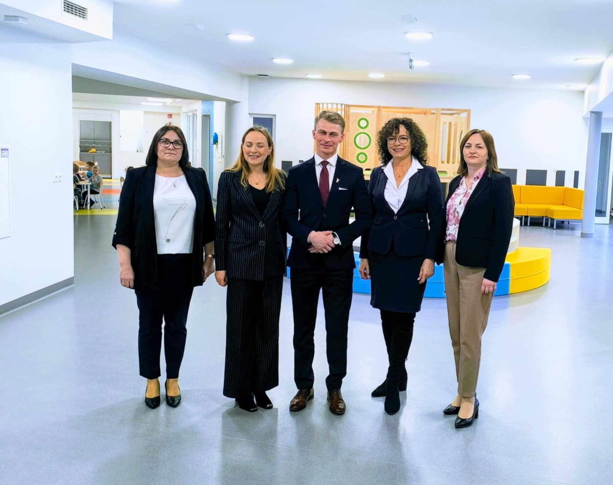 Pięć osób – cztery kobiety i jeden mężczyzna – stoi w jasnym, nowoczesnym wnętrzu, najprawdopodobniej w przedszkolu lub szkole. W tle widać kolorowe meble i dzieci siedzące przy stolikach. Wszyscy dorośli są elegancko ubrani, co sugeruje formalne spotkanie lub wizytę służbową.