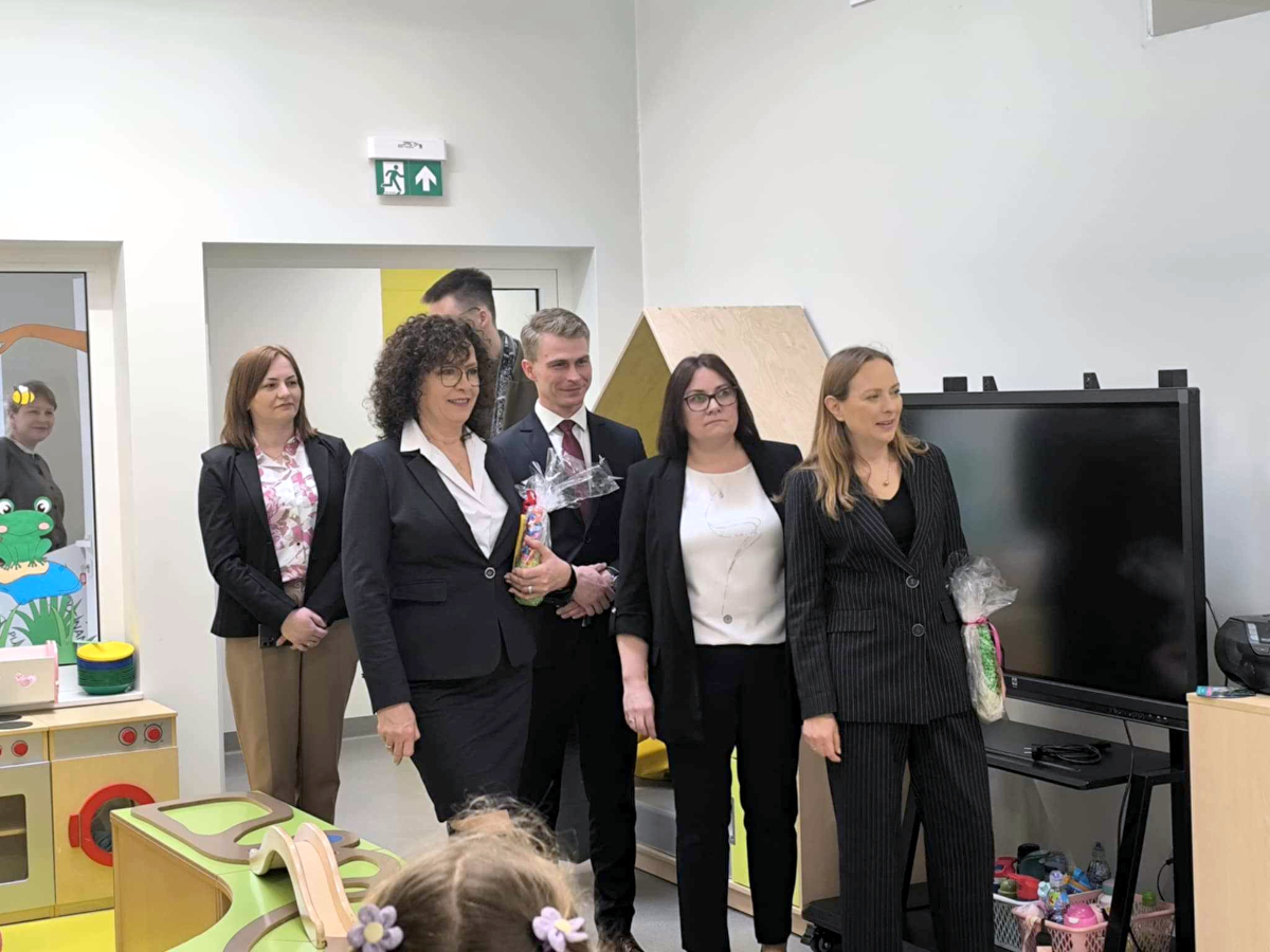 Grupa pięciu dorosłych – cztery kobiety i jeden mężczyzna – stoi w sali przedszkolnej, przy dużym ekranie i zabawkach. Dwie osoby trzymają upominki owinięte w celofan. W tle widać kolorowe meble i zabawki, a także dziecięcy kącik kuchenny. 