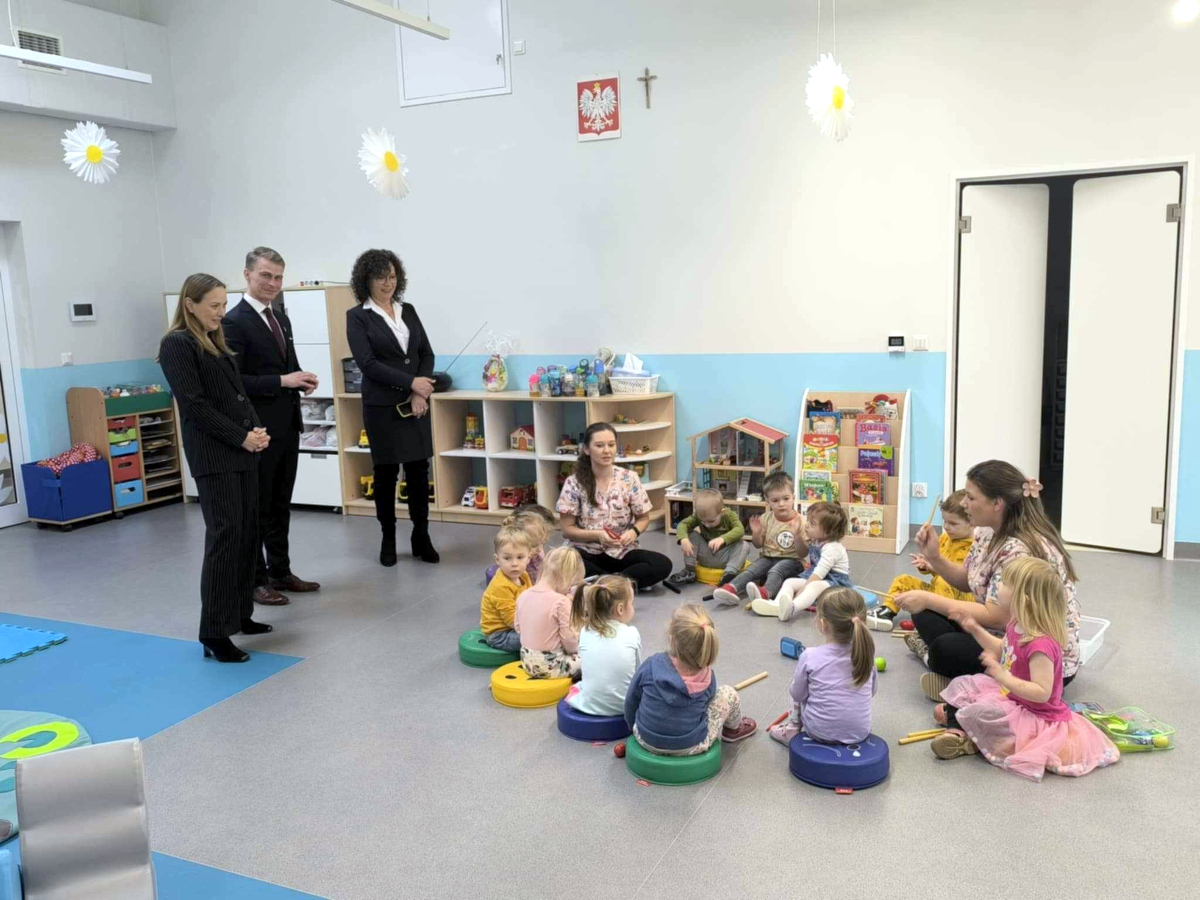 Grupa kilkunastu małych dzieci siedzi w kręgu na kolorowych poduszkach w sali przedszkolnej, uczestnicząc w zajęciach muzycznych z dwiema nauczycielkami. Troje dorosłych – dwie kobiety i mężczyzna w eleganckich strojach – stoi z boku i obserwuje zajęcia. W tle widoczne są półki z zabawkami i książkami, a na ścianie zawieszony jest godło Polski i krzyż.