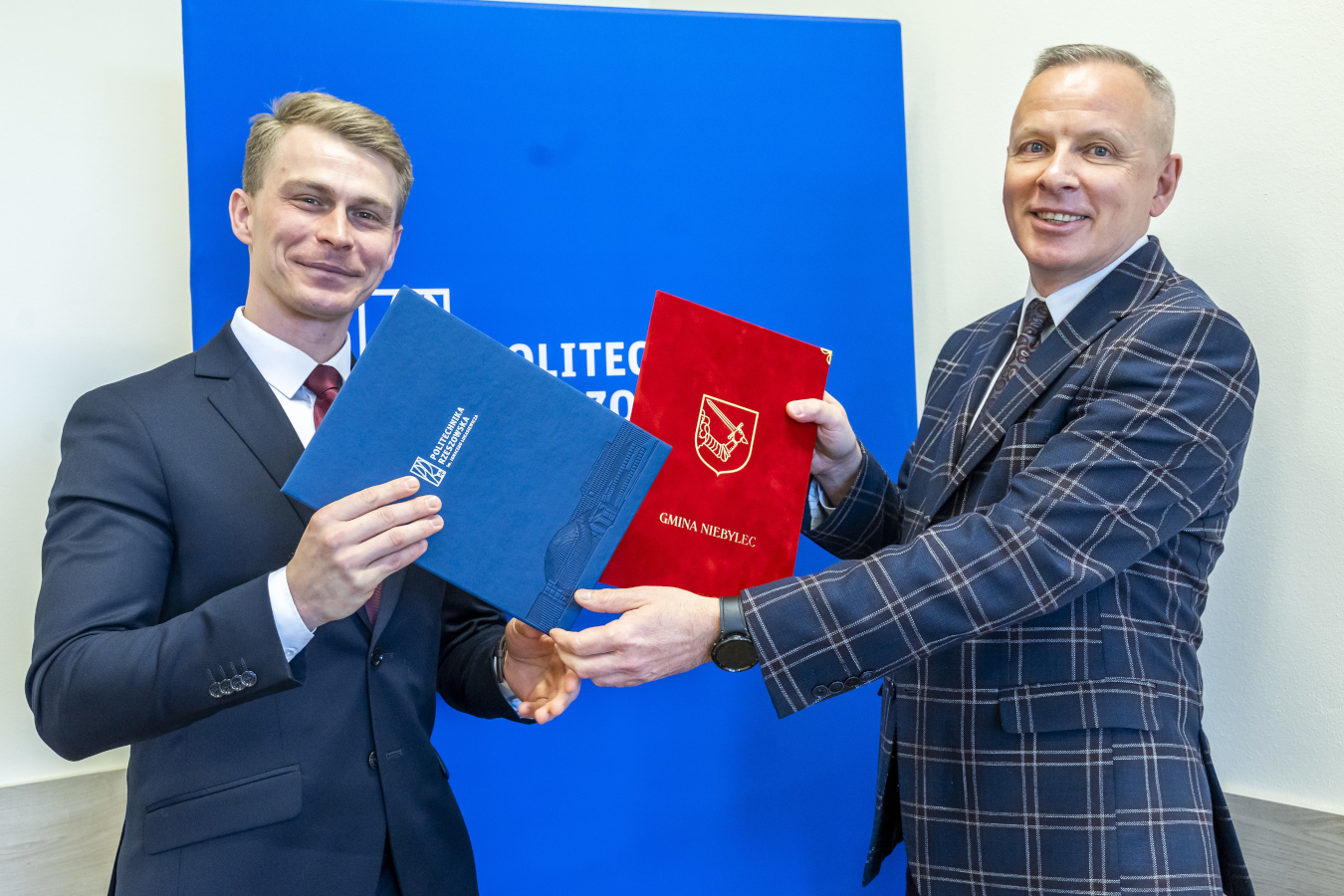Dwóch elegancko ubranych mężczyzn stoi obok siebie, uśmiechając się i trzymając w dłoniach dokumenty oprawione w okładki. Mężczyzna po lewej stronie trzyma niebieską okładkę z logo Politechniki Rzeszowskiej, a mężczyzna po prawej trzyma czerwoną okładkę z herbem Gminy Niebylec i napisem "GMINA NIEBYLEC". W tle widoczne jest niebieskie tło z częściowo widocznym białym napisem "POLITEC". Mężczyzna po lewej ma jasne włosy, białą koszulę i ciemny garnitur w prążki oraz czerwony krawat. Mężczyzna po prawej ma krótko ostrzyżone siwe włosy, białą koszulę, krawat w drobny wzór i ciemny garnitur w kratę. Obaj mężczyźni mają na lewych nadgarstkach zegarki.