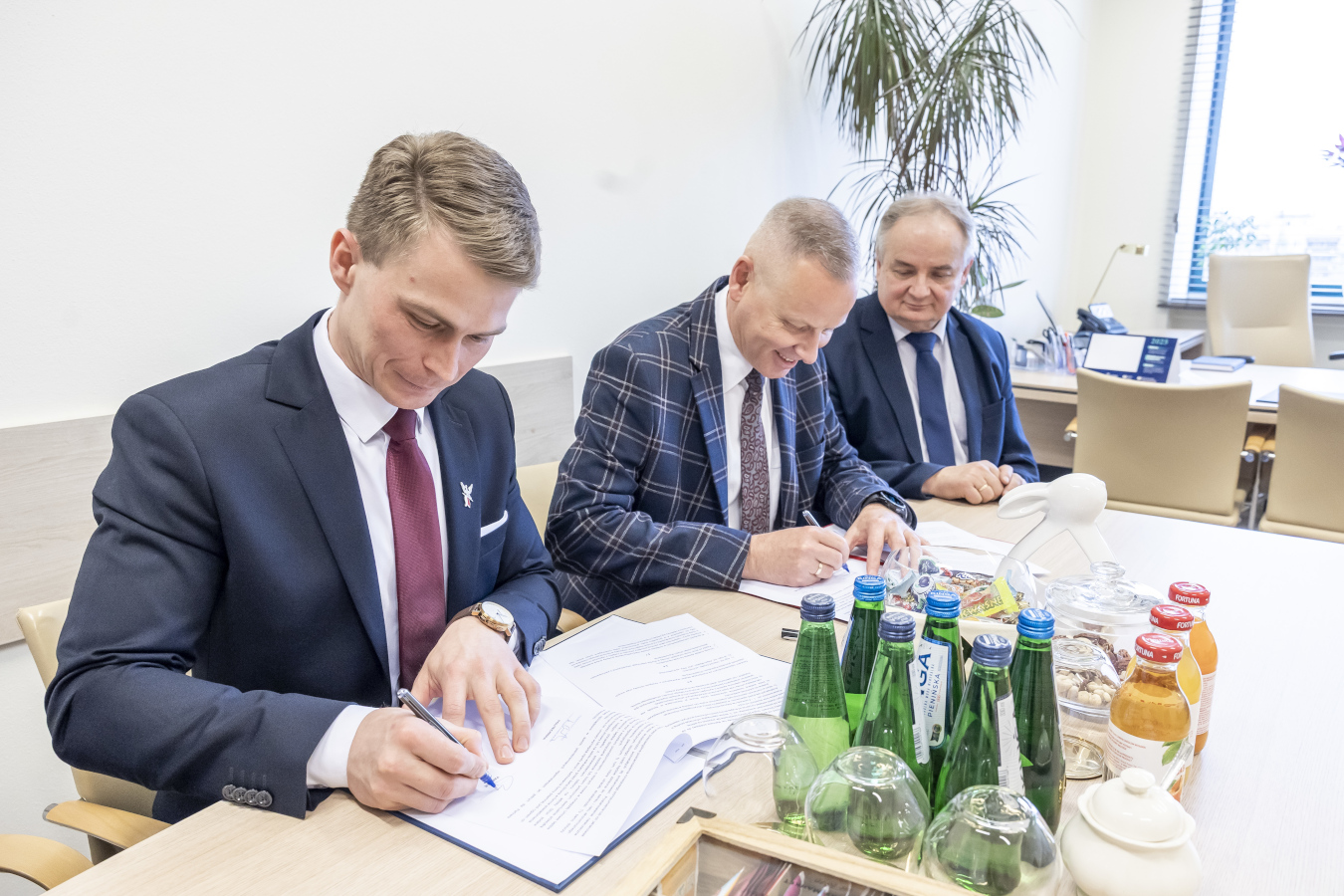 Opis zdjęcia: Trzech elegancko ubranych mężczyzn siedzi przy jasnym stole i podpisuje dokumenty. Mężczyzna po lewej stronie, ten sam co na poprzednim zdjęciu, pochyla się nad dokumentem i podpisuje go długopisem. Ma na sobie ciemny garnitur w prążki, białą koszulę i bordowy krawat. Mężczyzna w środku, również znany z poprzedniego zdjęcia, w niebiesko-szarym garniturze w kratę, białej koszuli i ciemnym krawacie w drobny wzór, również podpisuje dokument. Trzeci mężczyzna, siedzący po prawej stronie, w ciemnym garniturze, białej koszuli i niebieskim krawacie, przygląda się podpisywanym dokumentom z lekkim uśmiechem. Na stole przed nimi leżą rozłożone dokumenty, a także kilka butelek wody w zielonych etykietach i soku pomarańczowego. W tle widać jasne pomieszczenie z palmą w doniczce i innymi elementami wyposażenia biurowego. Scena sugeruje oficjalne podpisanie umowy lub porozumienia.