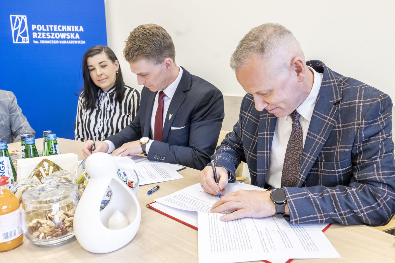 Trzy osoby siedzą przy jasnym stole i podpisują dokumenty. Mężczyzna po prawej stronie, w niebiesko-szarym garniturze w kratę i brązowym krawacie w drobny wzór, pochyla się nad dokumentem i podpisuje go czarnym długopisem. Obok niego siedzi mężczyzna w ciemnym garniturze, białej koszuli i bordowym krawacie, również podpisujący dokument. Między nimi siedzi kobieta w czarno-białej bluzce w paski, która przygląda się procesowi podpisywania. Na stole leżą dokumenty, butelki z wodą i sokiem pomarańczowym oraz biała dekoracyjna rzeźba. W tle widoczne jest niebieskie tło z białym napisem "POLITECHNIKA RZESZOWSKA im. Ignacego Łukasiewicza" i logo uczelni. Fragment osoby w szarym garniturze widoczny jest po lewej stronie kadru. Scena przedstawia formalne podpisanie umowy lub innego rodzaju dokumentu
