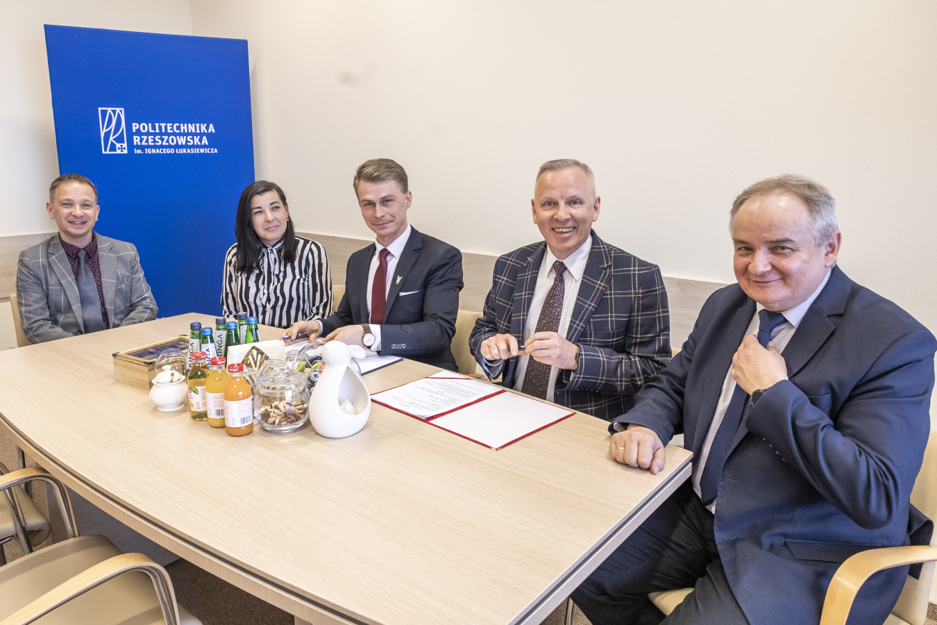 Pięciu elegancko ubranych mężczyzn i kobiet siedzi za jasnym, owalnym stołem w jasnym pomieszczeniu. W centrum stołu leżą otwarte dokumenty z czerwoną obwolutą. Po obu stronach dokumentów siedzą mężczyźni w garniturach, patrzący w stronę aparatu z uśmiechem. Mężczyzna po prawej ma na sobie ciemny garnitur w prążki i niebieski krawat w kropki, a mężczyzna po lewej ma ciemny garnitur, białą koszulę i bordowy krawat.  Po lewej stronie stołu siedzi kobieta w czarno-białej bluzce w paski, patrząca w stronę dokumentów. Obok niej siedzi mężczyzna w szarej marynarce w kratę i ciemnej koszuli. Na końcu stołu, po lewej stronie, siedzi mężczyzna w ciemnym swetrze i jasnej koszuli, również patrzący w stronę aparatu z uśmiechem.  Na stole, przed siedzącymi osobami, stoją butelki z wodą, sokami i przekąskami w szklanych naczyniach. W tle widoczna jest niebieska ściana z białym logo i napisem "POLITECHNIKA RZESZOWSKA im. Ignacego Łukasiewicza". Scena wygląda na spotkanie biznesowe lub uroczyste podpisanie dokumentów.
