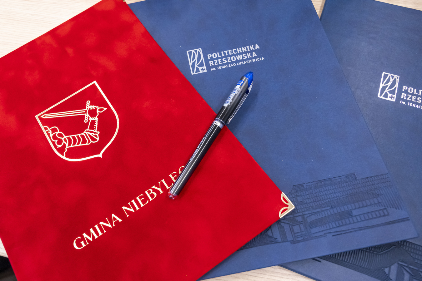 Na pierwszym planie leży czerwona teczka z herbem Gminy Niebylec w kolorze białym po lewej stronie oraz białym napisem "GMINA NIEBYLEC" biegnącym ukośnie w dół po prawej stronie. Na teczce spoczywa czarny długopis z przezroczystą niebieską zatyczką i srebrnymi elementami. W tle widoczne są dwie niebieskie teczki. Na jednej z nich, znajdującej się bliżej, widnieje białe logo Politechniki Rzeszowskiej oraz biały napis "POLITECHNIKA RZESZOWSKA im. Ignacego Łukasiewicza" ułożony w dwóch wierszach. Druga niebieska teczka, widoczna fragmentarycznie w prawym górnym rogu, ma identyczne logo i napis. Wszystkie trzy teczki leżą na jasnej, prawdopodobnie drewnianej powierzchni stołu.