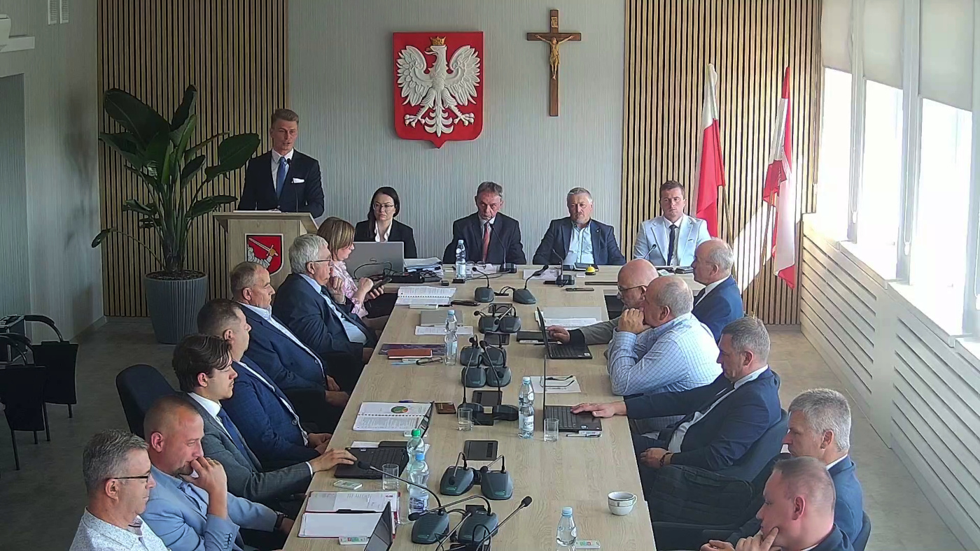Zdjęcie przedstawia sesję rady gminy lub powiatu. W jasnej sali konferencyjnej przy dużym stole obradowym siedzi około dwudziestu osób – głównie mężczyzn w garniturach. Przy pulpicie po lewej stronie stoi mężczyzna w ciemnym garniturze, który przemawia. Na ścianie za stołem widoczne są godło Polski (biały orzeł na czerwonym tle), krzyż oraz dwie flagi – polska i lokalna. Na stole znajdują się dokumenty, mikrofony, butelki z wodą i laptopy. W tle stoi duża doniczkowa roślina. Pomieszczenie jest dobrze oświetlone naturalnym światłem z okien po prawej stronie.