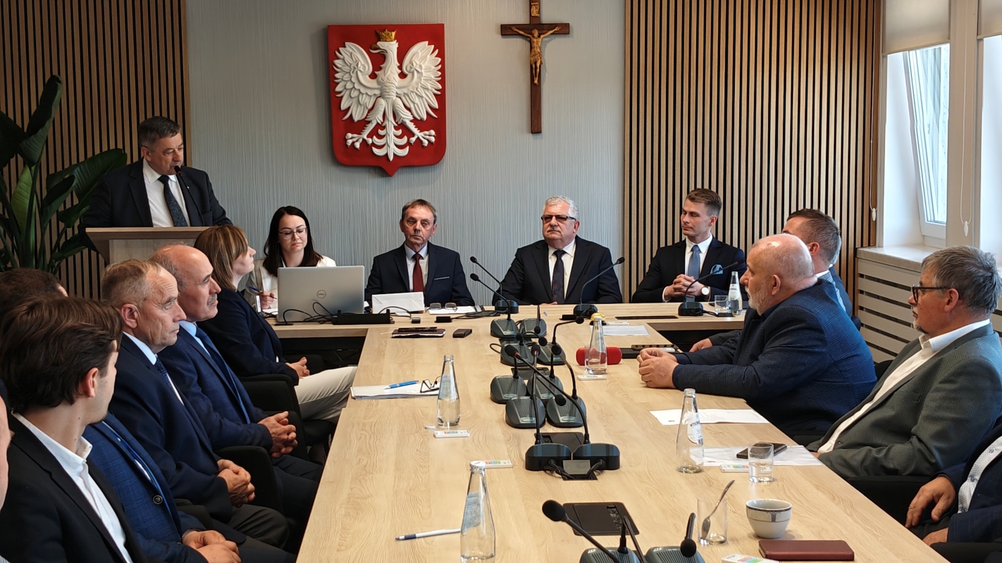 Sesja rady gminy w sali konferencyjnej z udziałem kilkunastu osób, głównie mężczyzn w garniturach. Jedna osoba stoi przy mównicy i przemawia, pozostałe siedzą wokół długiego stołu. Na stole znajdują się mikrofony, butelki z wodą, dokumenty i filiżanka. W tle widać godło Polski i krzyż, a po lewej stronie dużą roślinę doniczkową. Pomieszczenie jest jasne, z pionowymi drewnianymi panelami na ścianach i oknami po prawej stronie.