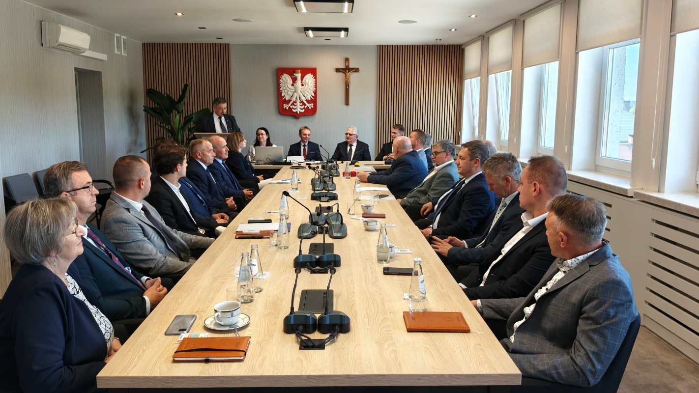 Zdjęcie przedstawia sesję rady gminy przy dużym, prostokątnym stole konferencyjnym. Wokół stołu siedzi kilkanaście osób, w tym kobiety i mężczyźni w garniturach i eleganckich ubraniach. Jedna osoba stoi przy mównicy, a dwie inne obsługują laptopy. Na ścianie w tle widoczne są godło Polski i krzyż. Na stole znajdują się mikrofony, butelki z wodą, filiżanki, notatniki i dokumenty. Pomieszczenie jest jasne, nowoczesne, z dużymi oknami po prawej stronie i drewnianymi panelami na końcu sali.