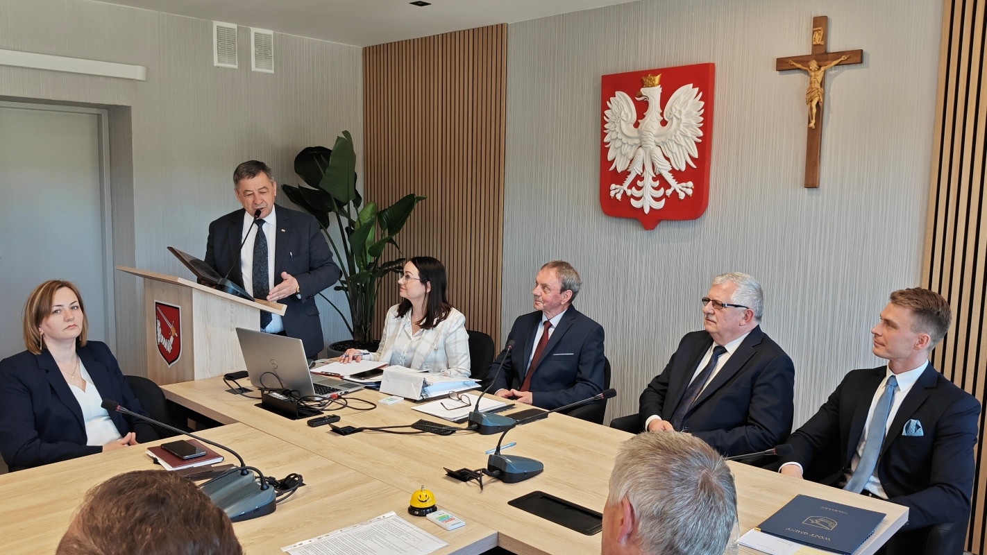 Mężczyzna w garniturze przemawia przy mównicy w sali konferencyjnej, obok godła Polski i krzyża. Przy stole siedzi sześć osób, w tym kobiety i mężczyźni w eleganckich strojach, niektórzy z laptopami i dokumentami. Na stole widać mikrofony, notatki, długopisy i żółty przycisk. W tle roślina doniczkowa i drewniane panele na ścianie.