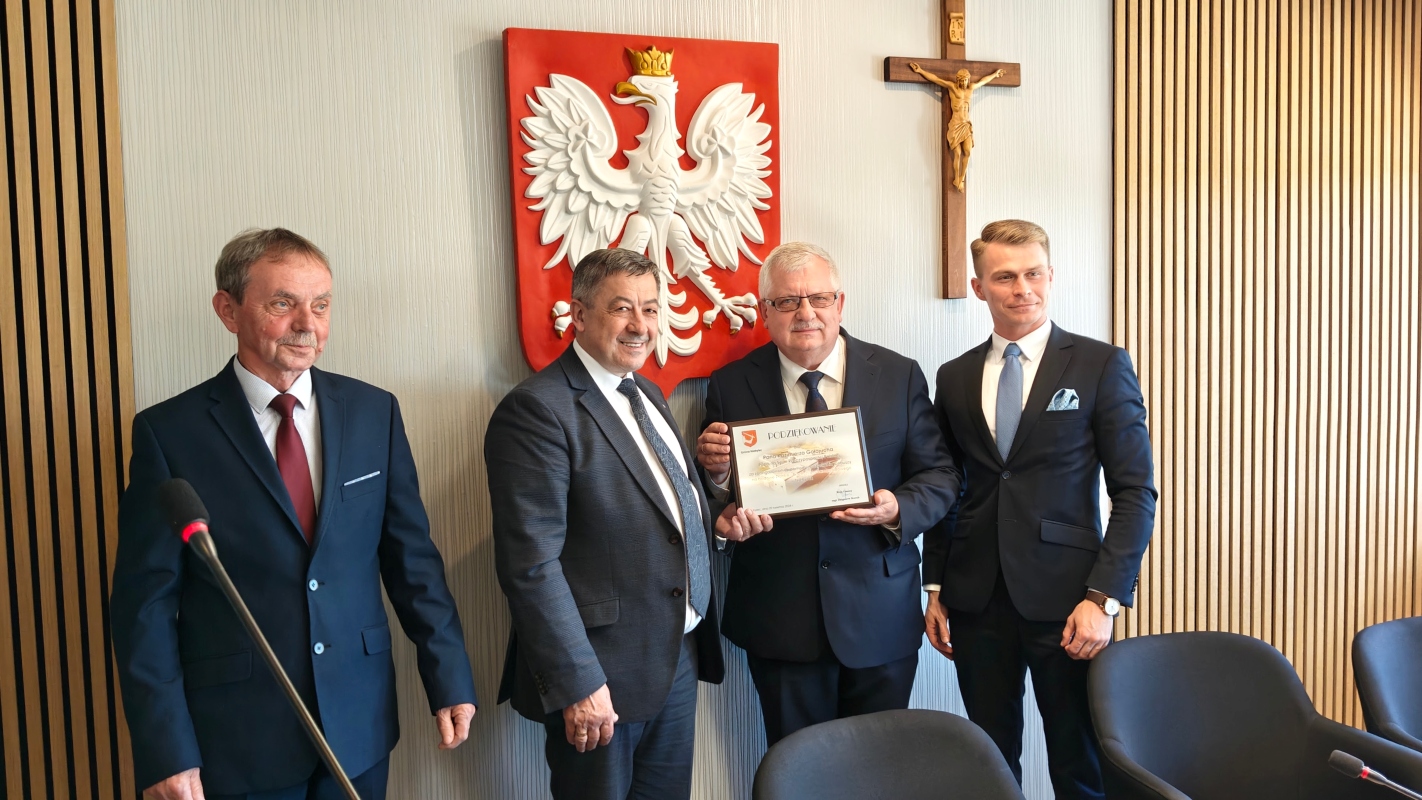 Czterech mężczyzn w garniturach pozuje do zdjęcia w sali urzędowej. Jeden z nich trzyma w rękach dyplom z napisem „Podziękowanie”. W tle widać godło Polski z białym orłem na czerwonym tle oraz krzyż z postacią Jezusa. Ściana ozdobiona jest pionowymi drewnianymi panelami.