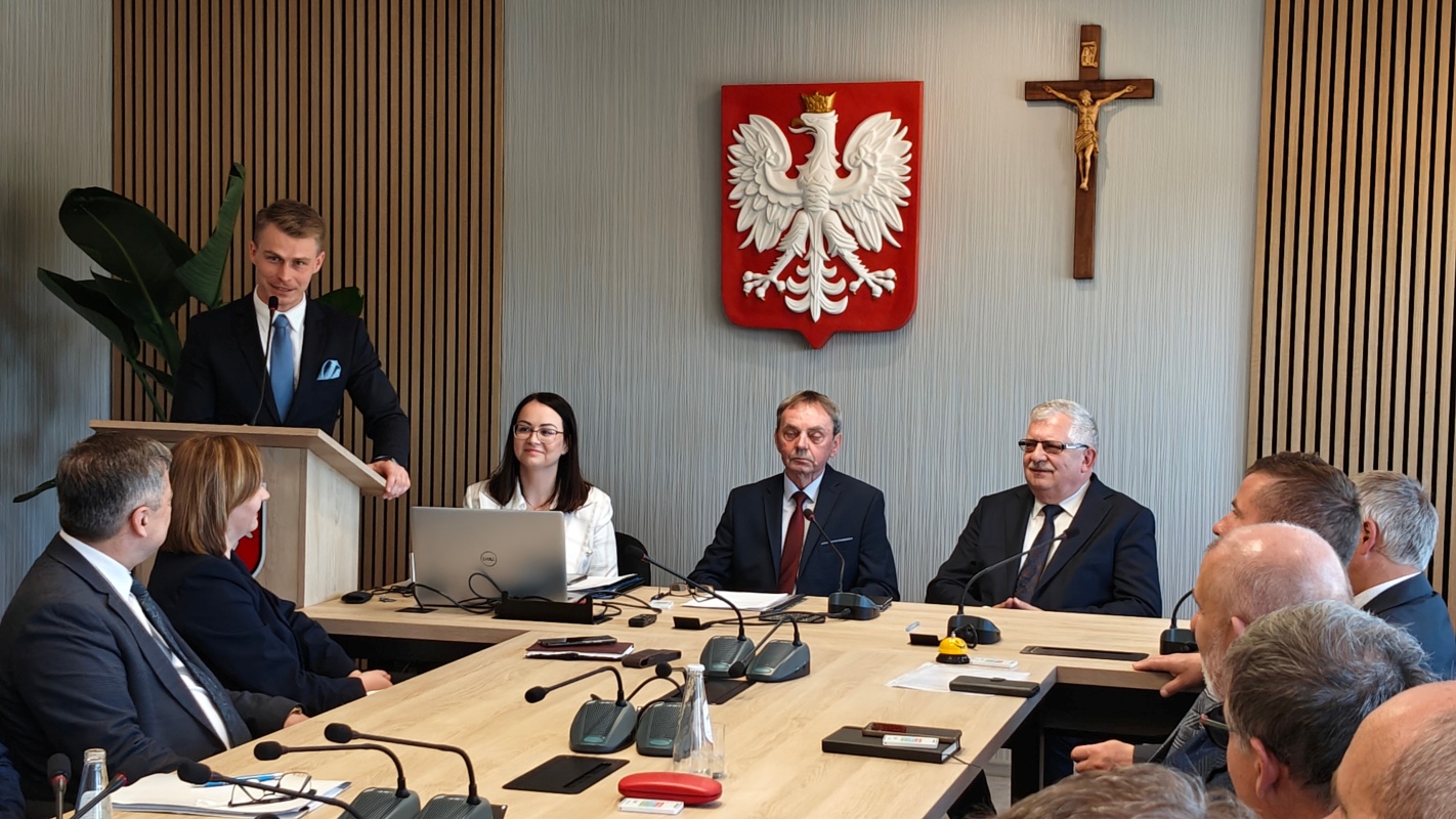 Mężczyzna w garniturze przemawia przy mównicy podczas oficjalnego spotkania w sali konferencyjnej. Przy stole siedzą uczestnicy – kobiety i mężczyźni w eleganckich strojach, niektórzy z laptopami i dokumentami. Na ścianie za nimi widoczne są godło Polski i krzyż z postacią Jezusa. Na stole znajdują się mikrofony, butelki z wodą, notatki i żółty przycisk. W tle roślina doniczkowa i drewniane panele ścienne.