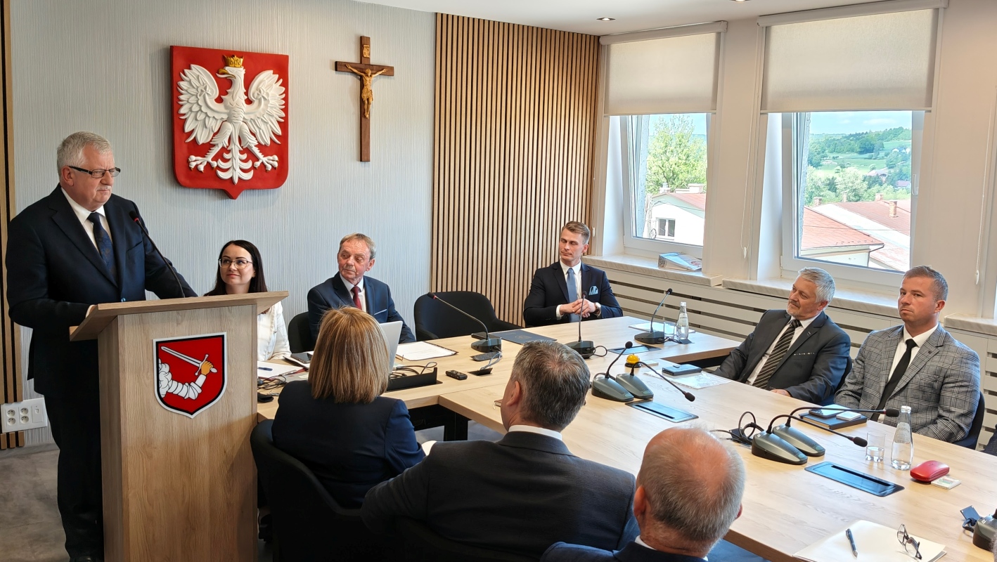 Mężczyzna w garniturze przemawia przy mównicy podczas oficjalnego spotkania w sali konferencyjnej. Uczestnicy, kobiety i mężczyźni w eleganckich strojach, siedzą wokół stołu i słuchają. Na ścianie za mówcą widoczne są godło Polski i krzyż. Na stole znajdują się mikrofony, dokumenty, butelki z wodą i filiżanki. Przez okna po prawej stronie widać dachy budynków i zielone wzgórza w tle. Pomieszczenie urządzone jest nowocześnie, z pionowymi drewnianymi panelami i jasnym wystrojem.