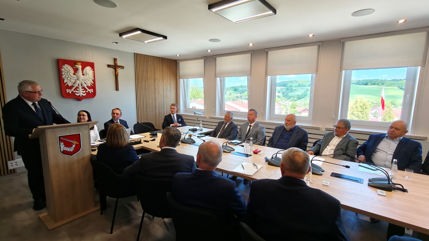 Zdjęcie przedstawia spotkanie w sali konferencyjnej. Mężczyzna w garniturze przemawia przy mównicy, na której widnieje herb z ramieniem trzymającym miecz. Przy stole siedzi kilkanaście osób w eleganckich strojach, słuchając uważnie. Na ścianie za mównicą widoczne są godło Polski i krzyż. Przez duże okna po prawej stronie widać zielony krajobraz, dachy budynków i powiewającą flagę Polski. Na stole znajdują się mikrofony, dokumenty, butelki z wodą i filiżanki. Pomieszczenie jest jasne, nowoczesne i przestronne.