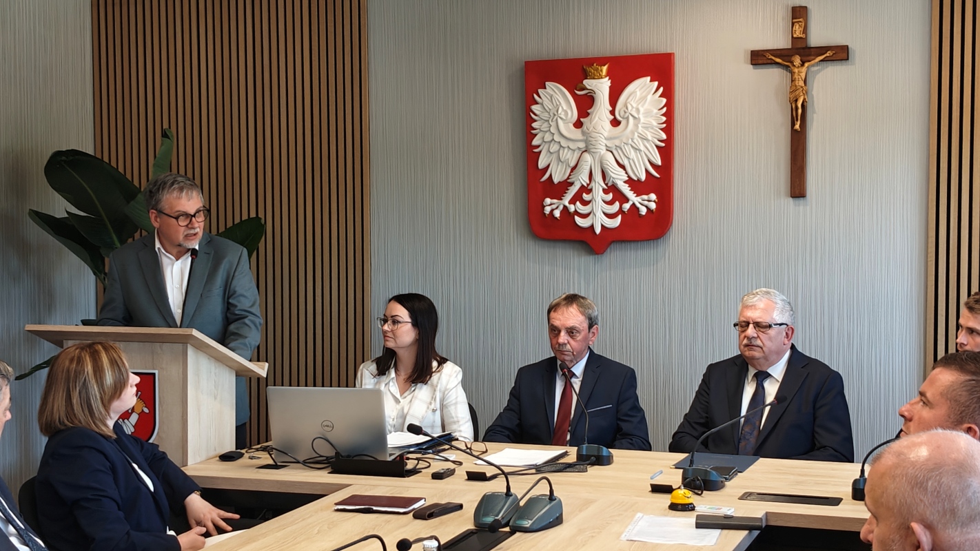 Mężczyzna w okularach i szarej marynarce przemawia przy mównicy podczas oficjalnego spotkania w sali konferencyjnej. Przy stole siedzą uczestnicy – kobiety i mężczyźni w garniturach – obok laptopa i dokumentów. Na ścianie za nimi widoczne są godło Polski i krzyż. Na stole znajdują się mikrofony, notatki, butelki z wodą i żółty przycisk. W tle pionowe drewniane panele i roślina doniczkowa.