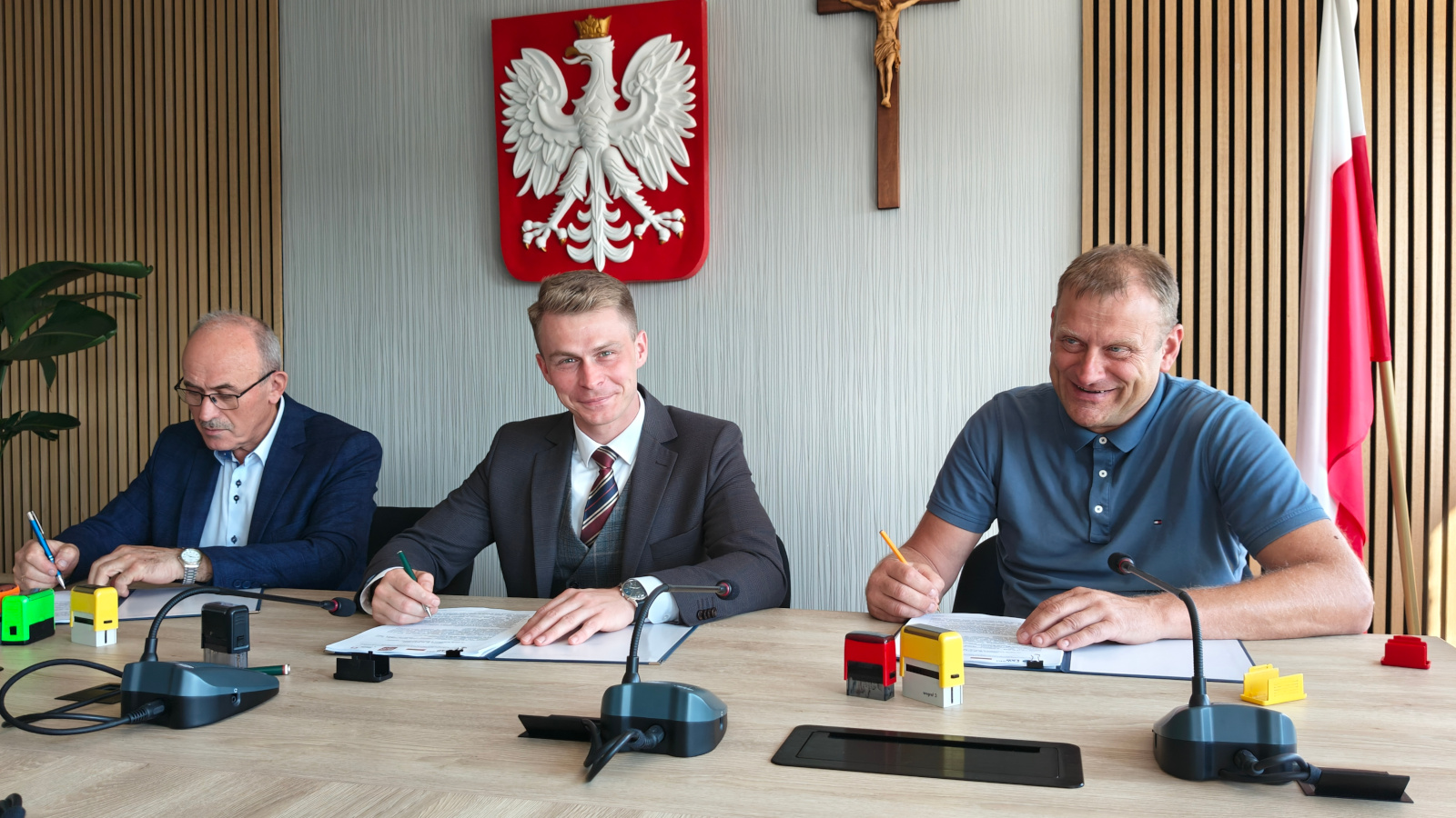 Trzech mężczyzn siedzących przy stole podpisuje dokumenty. W tle godło Polski, krzyż oraz flaga narodowa. Na stole leżą mikrofony, kolorowe pieczątki i dokumenty. Mężczyźni uśmiechają się i patrzą na dokumenty, jeden z nich ubrany w garnitur.