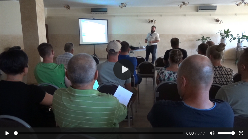 Opis alternatywny: Grupa ludzi siedzi tyłem do kamery w sali konferencyjnej, słuchając prezentacji. Na pierwszym planie widać mężczyzn w koszulkach, a z przodu sali stoi prowadzący z mikrofonem obok ekranu z rzutnikiem. Obraz przedstawia kadr z filmu, z widocznym przyciskiem odtwarzania i czasem trwania nagrania (3:17).
