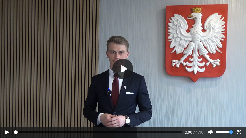 Mężczyzna w ciemnym garniturze, białej koszuli i bordowym krawacie stoi za ciemnym stołem. Ma jasną cerę i krótkie blond włosy. Jego dłonie są splecione przed nim. Za nim, na szarej ścianie, wisi godło Polski - biały orzeł w złotej koronie na czerwonym tle. Po lewej stronie mężczyzny widoczna jest ściana pokryta pionowymi, drewnianymi panelami. W dolnej części kadru znajduje się interfejs odtwarzacza wideo z przyciskiem pauzy, suwakiem postępu, licznikiem czasu "0:00 / 1:16" oraz ikonami głośności i ustawień.