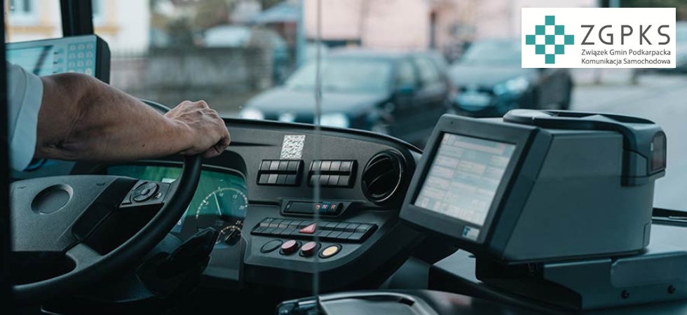Zdjęcie przedstawia miejsce pracy kierowcy autobusu miejskiego lub międzymiastowego. Po lewej stronie widać rękę kierowcy trzymającą kierownicę, a przez przednią szybę można dostrzec zaparkowane samochody i zabudowę mieszkalną, co sugeruje, że autobus stoi w ruchu miejskim lub na przystanku. Centralna część zdjęcia pokazuje pulpit z przyciskami i wskaźnikami – są one uporządkowane i nowoczesne, wskazując na nowszy model pojazdu. Po prawej stronie znajduje się duży terminal – prawdopodobnie urządzenie do sprzedaży biletów lub system kontroli biletów elektronicznych. Ekran terminala jest włączony i pokazuje interfejs z kilkoma liniami lub opcjami, ale nie da się dokładnie odczytać, co jest na nim wyświetlone. W prawym górnym rogu nałożone jest logo ZGPKS – Związku Gmin Podkarpacka Komunikacja Samochodowa – które symbolizuje organizację odpowiedzialną za transport publiczny w regionie Podkarpacia w Polsce.